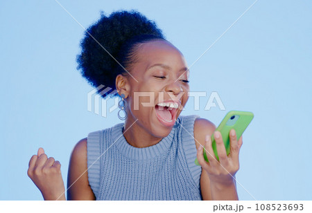Celebration. phone and young black woman in a studio with winning, achievement or success. Happy, smile and African female winner cheering with fist pump on cellphone isolated by blue background. Celebration. phone and young black woman in a studio with winning, achievement or success. Happy, smile and African female winner cheering with fist pump on cellphone isolated by blue background. 108523693