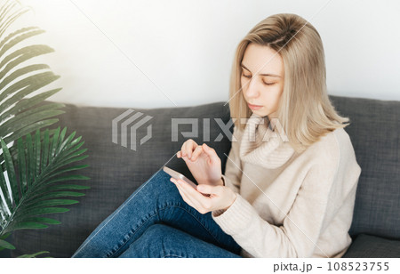 Happy young woman relaxing on comfortable couch, holding smartphone in hands. 108523755