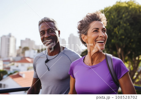 Adult international couple in sports outfits looking at camera with energetic cheerful smile. Happy loving mature man and woman jogging or exercising outdoors. Healthy lifestyle in urban environment. 108528702