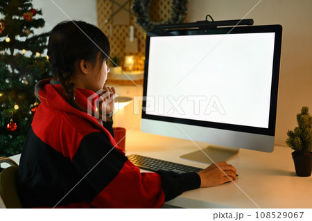 Side view of young woman in sweater using computer near decorated Christmas tree in cozy living room Side view of young woman in sweater using computer near decorated Christmas tree in cozy living room 108529067