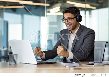 A man at the workplace inside the office is watching a sports match, a businessman in headphones and with a laptop is cheering at the workplace, a joyful boss, celebrating a victory, smiling. 108529463