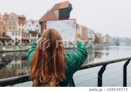 Attractive young female tourist is exploring new city. Redhead girl with backpack holding a paper map on city street in Gdansk. Traveling Europe in autumn. Famous Zuraw crane, Motlawa river Attractive young female tourist is exploring new city. Redhead girl with backpack holding a paper map on city street in Gdansk. Traveling Europe in autumn. Famous Zuraw crane, Motlawa river 108530573