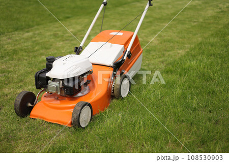 Top view of modern orange-grey electric lawn mower cutting bright lush green grass. Gardening work tools. Rotary lawn mower machine on lawn. Professional lawn care service. Place for text 108530903
