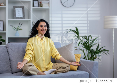 Young beautiful woman meditating at home sitting on sofa in living room in lotus pose, latin american joyful smiling, dreaming and visualizing future achievement results and plans. Young beautiful woman meditating at home sitting on sofa in living room in lotus pose, latin american joyful smiling, dreaming and visualizing future achievement results and plans. 108535141