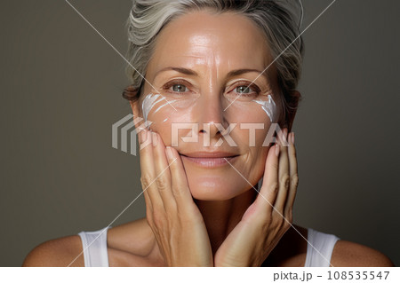 Close-up of middle-aged Caucasian woman touching her face to apply moisturizer. Smiling face of adult grey-haired lady with daily cream, facial cosmetics. Skin care. Grey background, copy space. 108535547
