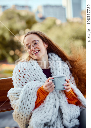 Portrait of happy smiling young girl with long hair sits covered blanket and drinks warming tea on bench in park. Autumn mood. 108537789