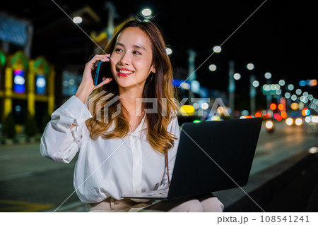 Woman is immersed in her work on a bustling city street corner at night, multitasking with her laptop and cell phone and utilizing convenience and versatility of modern technology to stay productive. Woman is immersed in her work on a bustling city street corner at night, multitasking with her laptop and cell phone and utilizing convenience and versatility of modern technology to stay productive. 108541241
