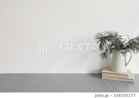 Elegant Christmas interior, home office. Ceramic vase, jug with green pine tree branches on old books. Winter home decor. Empty white wall background mockup. Grey table, desk. No people. Elegant Christmas interior, home office. Ceramic vase, jug with green pine tree branches on old books. Winter home decor. Empty white wall background mockup. Grey table, desk. No people. 108550177