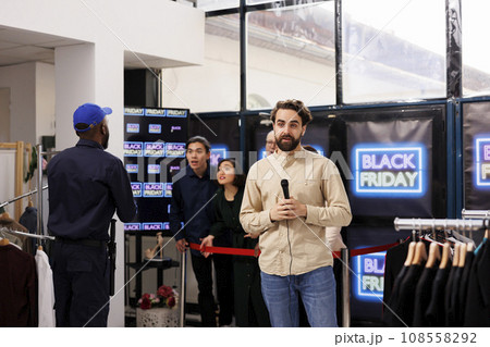 Young man TV reporter covering news real-time, live streaming during Black Friday sales. Correspondent standing at store entrance talking into microphone making live newscast about shopping madness Young man TV reporter covering news real-time, live streaming during Black Friday sales. Correspondent standing at store entrance talking into microphone making live newscast about shopping madness 108558292
