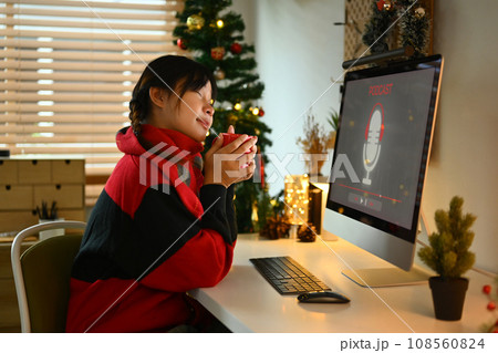 Happy young woman drinking hot chocolate and listening to podcast or radio playlist while resting on Christmas eve at home Happy young woman drinking hot chocolate and listening to podcast or radio playlist while resting on Christmas eve at home 108560824