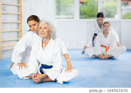 Elderly woman in kimono performing butterfly stretch with help of female coach Elderly woman in kimono performing butterfly stretch with help of female coach 108561670