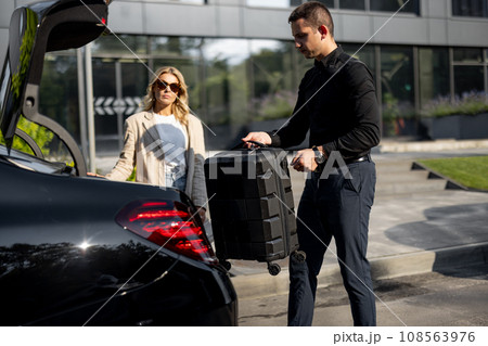 Businesswoman waits while chauffeur packs a suitcase in car trunk 108563976