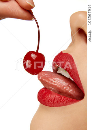 Cropped photo. Close up of woman with red lips licking sweet red cherry. Cherries in woman mouth. Summer sexy fruits. 108567916