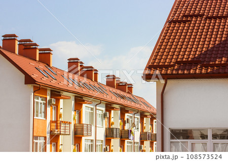 Facade of new white apartment building with red terra cotta tile roof against blue sky. Newly built modern comfortable residential house balcony. Real estate investment. Chimney, small window in roof. 108573524