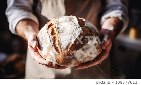 A man baker in a gray apron stands against a dark background and holds bread 108577613