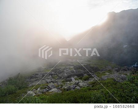 Colorful green landscape with stones on the hill on background of giant mountain wall in misty sunlight. Minimalist vivid sunny scenery with sun and solar flare. Minimal alpine view. Scenic minimalism 108582668