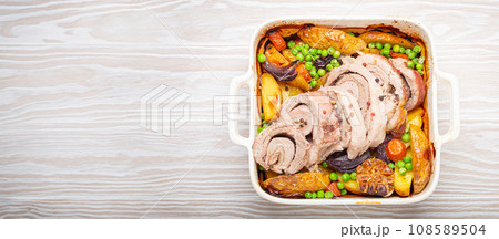 Rolled sliced pork roasted in white casserole dish with potatoes, vegetables and herbs on rustic white wooden background top view. Baked pork roll with vegetables for dinner Rolled sliced pork roasted in white casserole dish with potatoes, vegetables and herbs on rustic white wooden background top view. Baked pork roll with vegetables for dinner 108589504