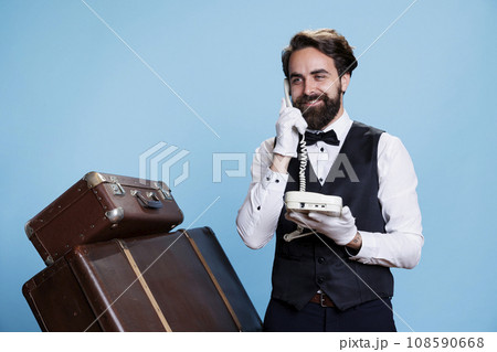 Guests call at hotel reception on landline phone, bellhop offering his excellent concierge services through wired telephone. Experienced classy bellboy talks to people against blue background. Guests call at hotel reception on landline phone, bellhop offering his excellent concierge services through wired telephone. Experienced classy bellboy talks to people against blue background. 108590668