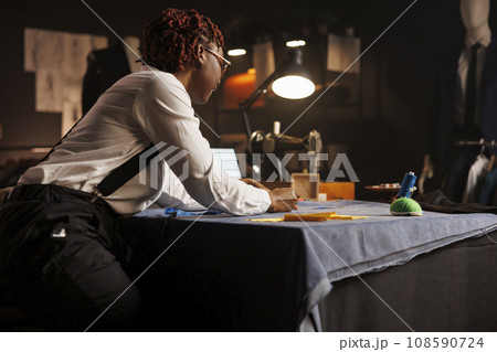 African american suitmaker doing fabric measurements in professional tailoring studio. Expert dressmaker working on manufacturing custom made stylish sartorial outfit for client 108590724