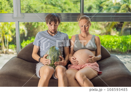 A humorous image: a pregnant woman and her husband playfully use a watermelon in place of a belly, comically highlighting the challenges of navigating with a pregnant bump A humorous image: a pregnant woman and her husband playfully use a watermelon in place of a belly, comically highlighting the challenges of navigating with a pregnant bump 108593485
