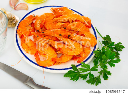 Plate with raw red shrimps, greens, condiments and knife on kitchen table Plate with raw red shrimps, greens, condiments and knife on kitchen table 108594247
