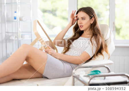 Satisfied young woman patient holding mirror lying on clinical chair 108594352
