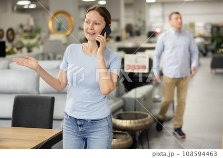 Happy female customer in furniture store talks on phone and consults with spouse about purchase Happy female customer in furniture store talks on phone and consults with spouse about purchase 108594463