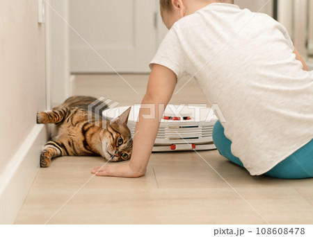 A little boy and a beautiful domestic leopard cat of the Bengal breed have fun playing in a home interior with a robot vacuum cleaner that cleans the apartment. 108608478