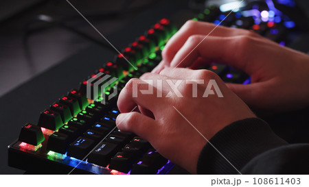 Male hands is typing on the computer keyboard, close up view. Man working on the pc computer. Human hands typing on the colourful gaming keyboard. Fingers typing on the led rgb keyboard. 108611403