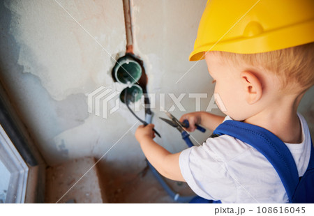 Close up of baby electrician cutting wires with pliers in building under construction. Kid in safety helmet fixing cables in wall while working on home renovation. Close up of baby electrician cutting wires with pliers in building under construction. Kid in safety helmet fixing cables in wall while working on home renovation. 108616045