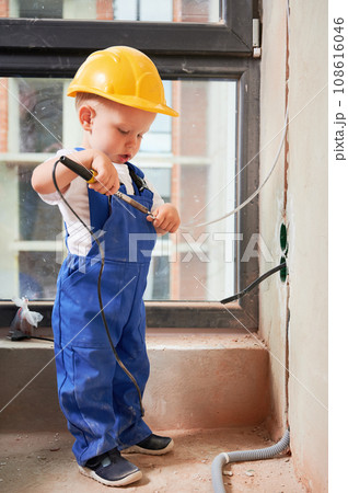 Kid welding electrical wire with electric soldering iron. Child electrician in work overalls and safety helmet installing electrical cables and sockets in apartment under renovation. 108616046