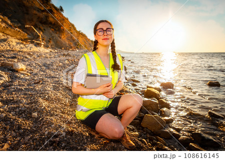 Earth day. Smiling woman wearing vest and holds electronic tablet squatting on beach near sea. Concept of ocean's pollution and protection of environment Earth day. Smiling woman wearing vest and holds electronic tablet squatting on beach near sea. Concept of ocean's pollution and protection of environment 108616145