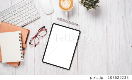 White wooden desk with blank screen tablet, keyboard, mouse, eyeglass, notebook, pen and cup of coffee, Top view flat lay with copy space. 108629238
