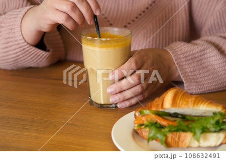 Woman in a Cosy Warm Sweater Holding a Cup of Coffee. Healthy Turmeric Autumnal Spicy Beverage. Pumpkin Latte with Curcuma in Female Hands Close up. Immunity Boosting Hot Drink of Fall Season. Cropped 108632491
