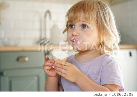 Baby girl enjoying ice cream. Pretty little toddler eating an ice-cream indoors, at home. Dining room background. Small child eats plombir and cream messy on her mouth. Cute kid with tasty sweet food. Baby girl enjoying ice cream. Pretty little toddler eating an ice-cream indoors, at home. Dining room background. Small child eats plombir and cream messy on her mouth. Cute kid with tasty sweet food. 108632508