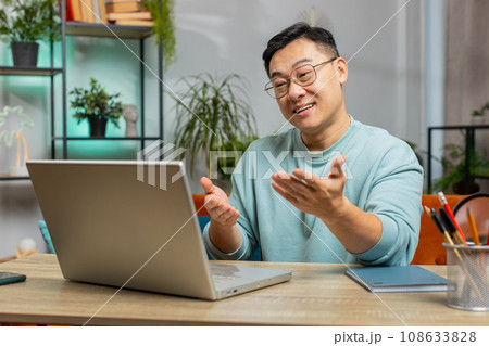 Asian man sitting on home couch, looking at laptop, making video conference call with friends family 108633828