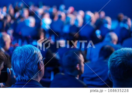 Businessmen listening to speaker giving presentation in illuminated auditorium at export forum  108635228