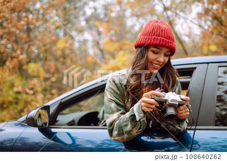 Beautiful woman takes pictures on camera from the car window. Smiling woman enjoying autumn weather. Beautiful woman takes pictures on camera from the car window. Smiling woman enjoying autumn weather. 108640262
