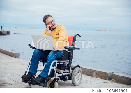 A man with disabilities sits with a laptop by the sea A man with disabilities sits with a laptop by the sea 108641901