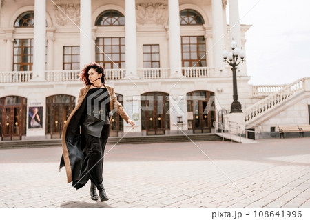 Woman street lifestyle. Image of stylish woman walking through European city on sunny day. Pretty woman with dark flowing hair, dressed in a beige raincoat and black, walks along the building. 108641996