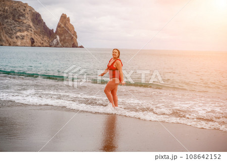 Woman in a bathing suit at the sea. A fat young woman in a red swimsuit enters the water during the surf Woman in a bathing suit at the sea. A fat young woman in a red swimsuit enters the water during the surf 108642152