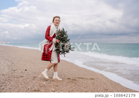 Blond woman holding Christmas tree by the sea. Christmas portrait of a happy woman walking along the beach and holding a Christmas tree in her hands. Dressed in a red coat, white dress. Blond woman holding Christmas tree by the sea. Christmas portrait of a happy woman walking along the beach and holding a Christmas tree in her hands. Dressed in a red coat, white dress. 108642179