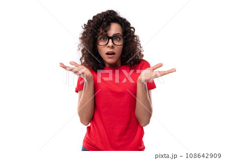 young surprised curly brunette woman dressed in a red t-shirt on a white background with copy space 108642909