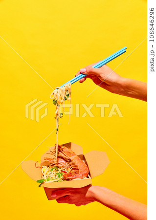Unrecognizable woman eating delicious hot WOK with shrimps with chopstick from paper box isolated yellow studio background 108646296