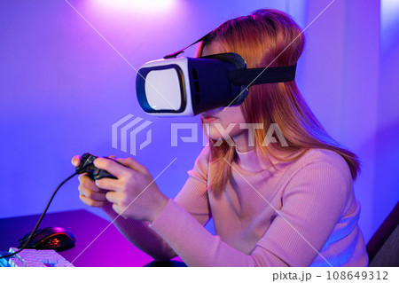 Gamer in VR headset glasses exploring metaverse play video game online with joystick on computer neon lights, Excited woman virtual playing gamepad controller she very determined at home 108649312
