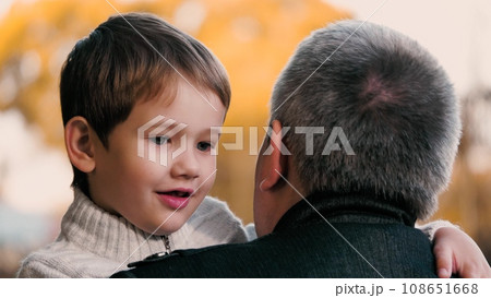 Father holds smiling son in arms with warmth while stands in park. Father amid ambiance of park holds son creating moment. Father in setting of park holding son in arms underscoring beauty of season Father holds smiling son in arms with warmth while stands in park. Father amid ambiance of park holds son creating moment. Father in setting of park holding son in arms underscoring beauty of season 108651668