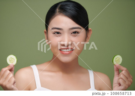 Young beautiful asian lady holding slices of cucumber near her face and smiling Young beautiful asian lady holding slices of cucumber near her face and smiling 108658713