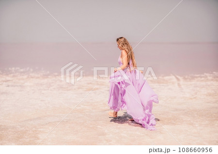 Woman pink salt lake. Against the backdrop of a pink salt lake, a woman in a long pink dress takes a leisurely stroll along the white, salty shore, capturing a wanderlust moment. 108660956