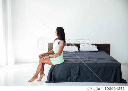 Brunette woman sitting on bed in bright room in bedroom Brunette woman sitting on bed in bright room in bedroom 108664851