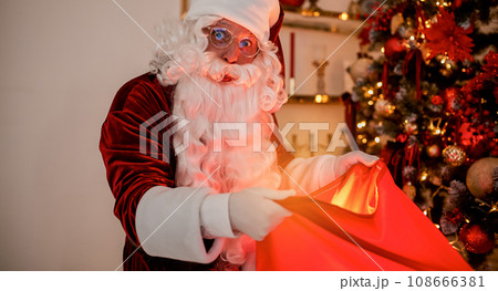 Surprised Santa Claus with glowing gift near christmas tree. New year and Merry Christmas concept Surprised Santa Claus with glowing gift near christmas tree. New year and Merry Christmas concept 108666381